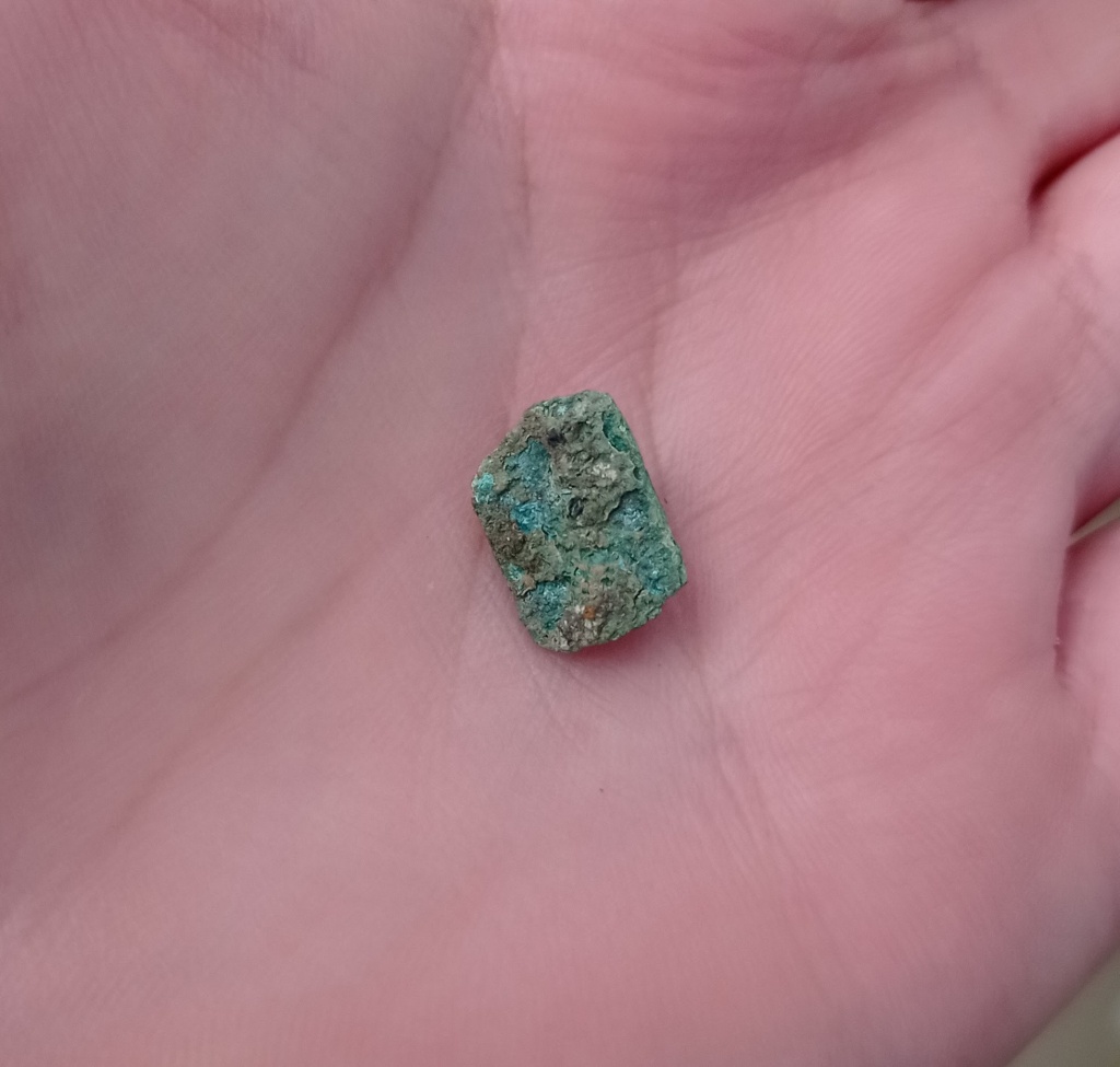 Corroded copper alloy fitting sits in a hand. the corrosion is light green and hints of aquamarine, which are minerals formed in the corrosion process.
