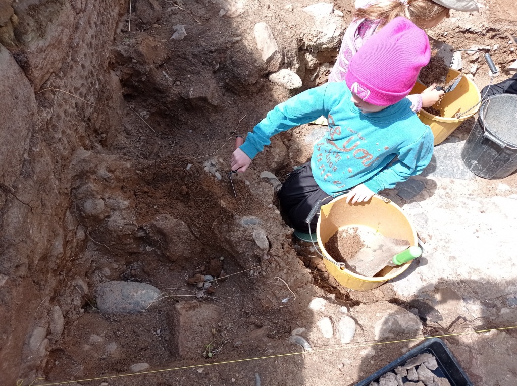 A young girl in a pink beanie and blue hoodie uses her pointing trowel to carefully excavate cobbles from an elevated ridge.