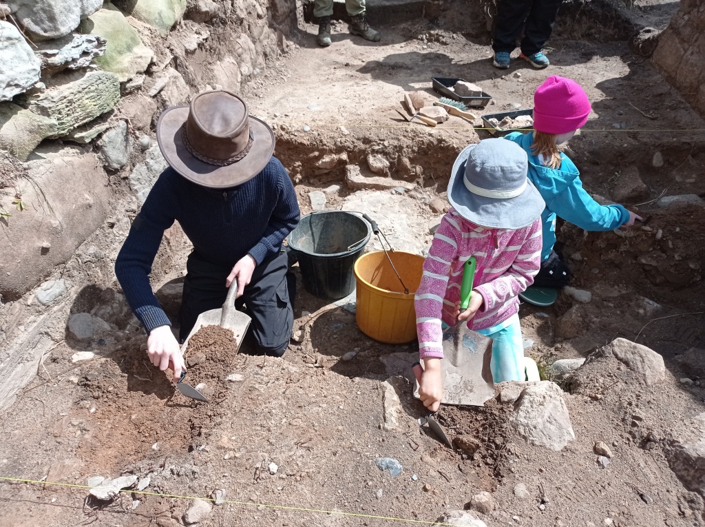 Three children in hats kneel in a small archaeological trench, scooping loose soil into their shovels to put in the black and yellow buckets behind them.