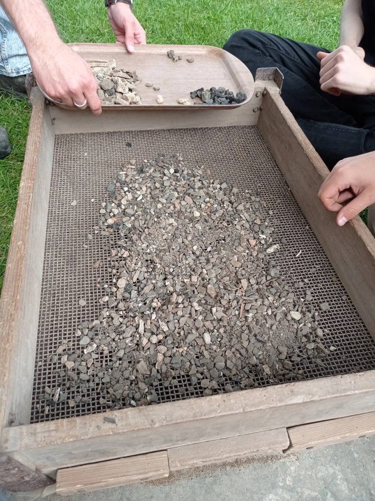 Multiple hands rest near a wooden-edged sieve, preparing to sort through a mass of light pinkish grey gravel. A pair of hands rests a wooden tray with sorted piles of artefacts including animal bone, mortar, charcoal, and slag.