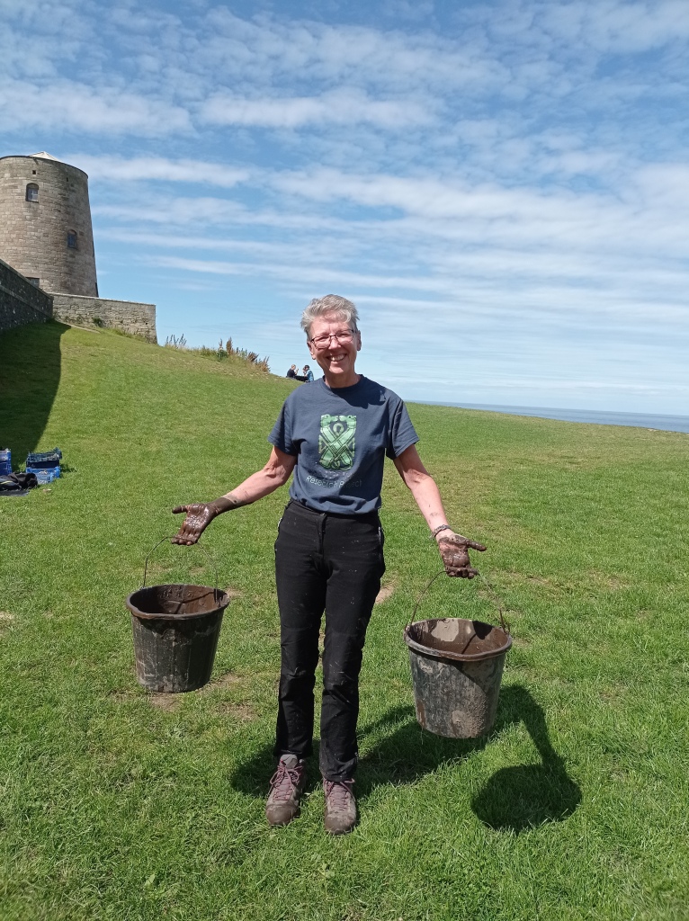 A woman in a BRP t-shirt carries buckets of silt away across the West Ward grass. A blue sky with linear clouds is behind her. Her hands are covered in mud up to her wrists, but she's smiling.