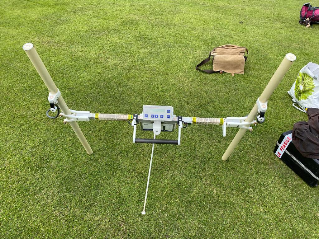 H-shaped rig standing upright on grass, with a light brown messenger bag in the background and a black hard briefcase on the right. 