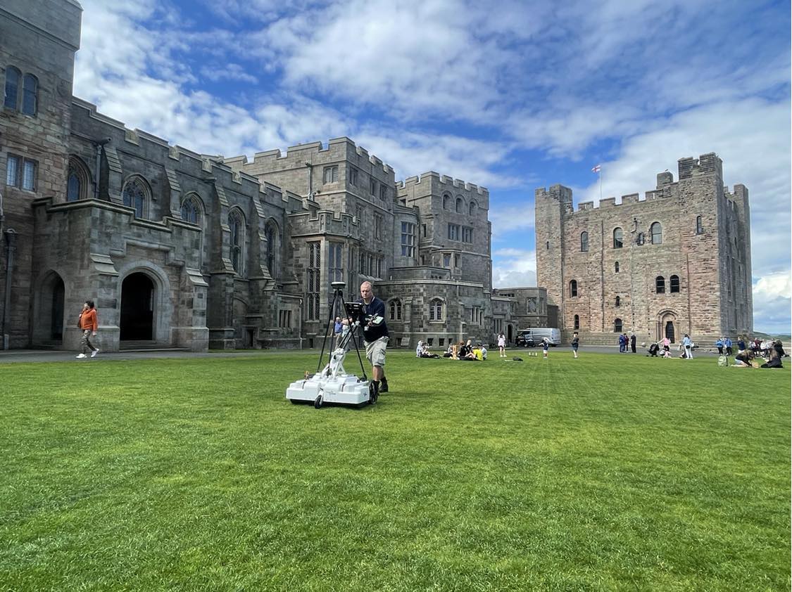 A man pushes a white cart that looks like a lawn mower with a tripod on it across impeccably mowed grass in the Inner Ward. Behind him stands pseudo-gothic buildings and a Norman square keep.