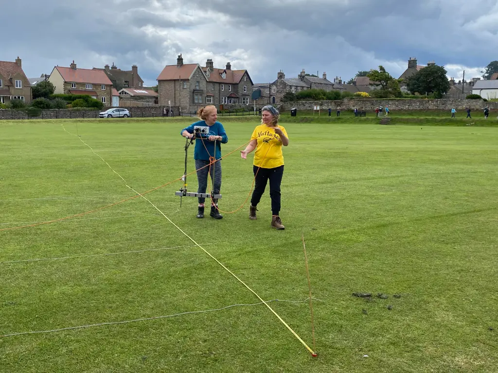 Two women walk across the green along a surveyor's tape laughing holding a resistivity array.