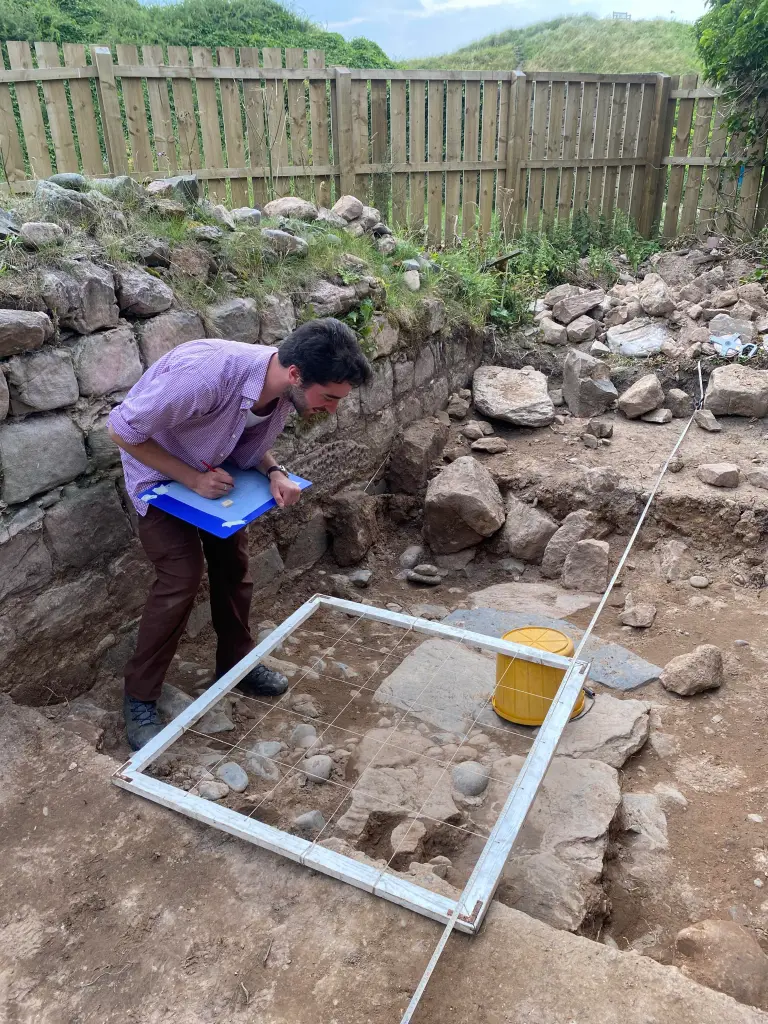 A young man leans over a drawing board in a trench next to a stone wall. He looks at a white frame with string to help copy what he's seeing in the ground to his record sheet.