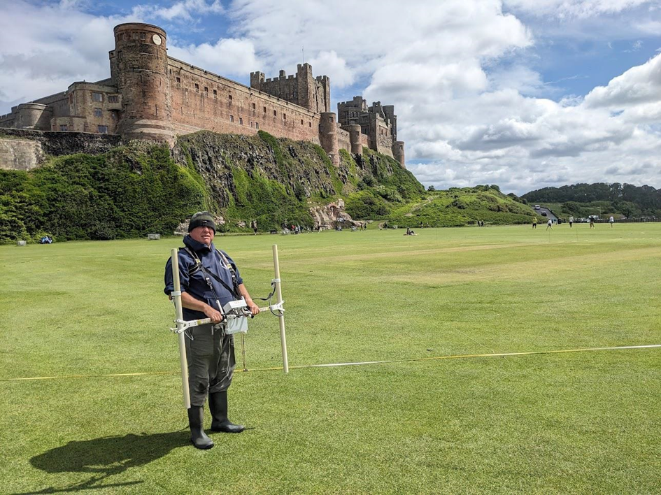 Person stood on large grassy area in front of large castle situated on rock promontory. Man is holding a white twin probe device with computer attached in middle