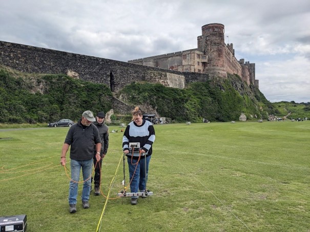 Three people stood on large grassy area in front of large castle situated on rock promontory. Person is holding a white twin probe device with computer attached in middle with wires hanging down